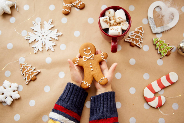 Vánoční domácí perníkové sušenky v dětských rukách mezi slavnostní vánoční ozdoby s teplým nápojem a marsmallow. Estetická vánoční atmosféra, domácí útulnost a vřelý koncept - Fotografie, Obrázek