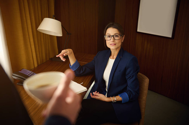Sukces kobiety szefa, biznesmenki siedzącej przy stole w pokoju hotelowym w świetle lampy stołowej i dyskutującej ze swoim kolegą partnerem biznesowym trzymającym zamazaną filiżankę kawy na pierwszym planie - Zdjęcie, obraz