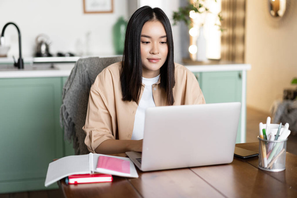 Azjatka studiująca w domu, koncepcja kształcenia na odległość. Kobieta uczy się z laptopem. Młoda kobieta pracuje zdalnie, siedzi w kuchni z książkami i komputerem - Zdjęcie, obraz