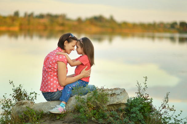 Pikkutyttö halaa äitiään järven rannalla.
 - Valokuva, kuva