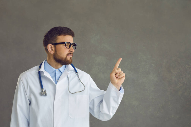 Handsome young doctor isolated on gray background points finger at copy space for text - Foto, Bild