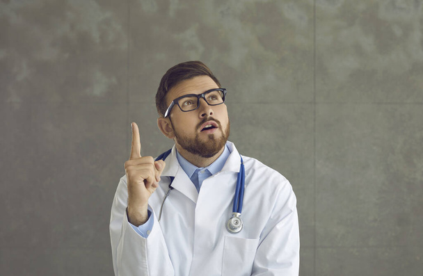 Young doctor in glasses and lab coat with stethoscope thinking with one finger pointing up - Foto, Bild