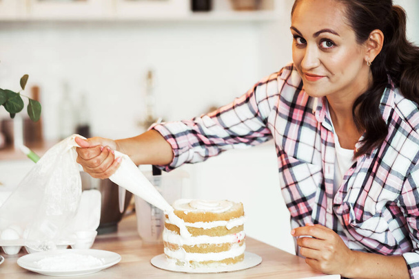 Beautiful pleased pastry chef woman making cake with cream at cozy kitchen - 写真・画像