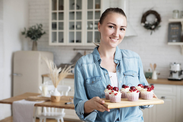 Work in confectionery workshop. Handmade cupcakes. Smiling pretty young woman pastry chef, demonstrating tasty fresh cupcales with berries to camera - Zdjęcie, obraz