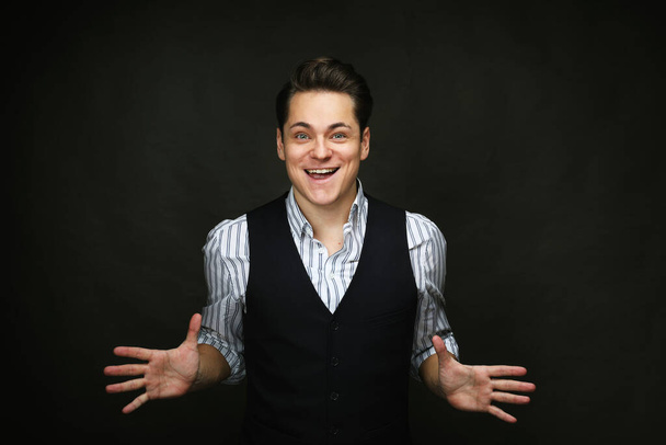 Young man wearing vest and shirt looking at the camera smiling with open arms for hug over black background. Cheerful expression embracing happiness. - Fotoğraf, Görsel