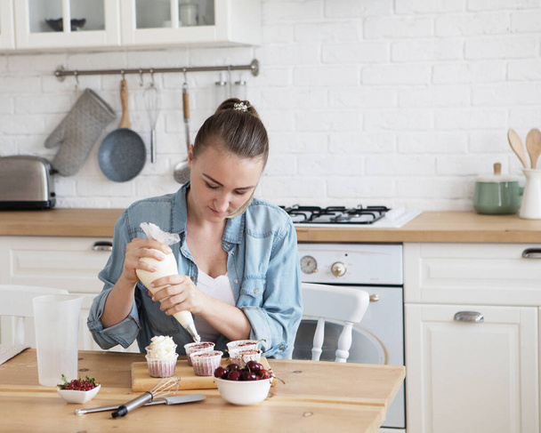 Cooking, Home Business, Freelance, People and Hobbies concept: A young woman wearing casual decorates muffins with white cream. - 写真・画像