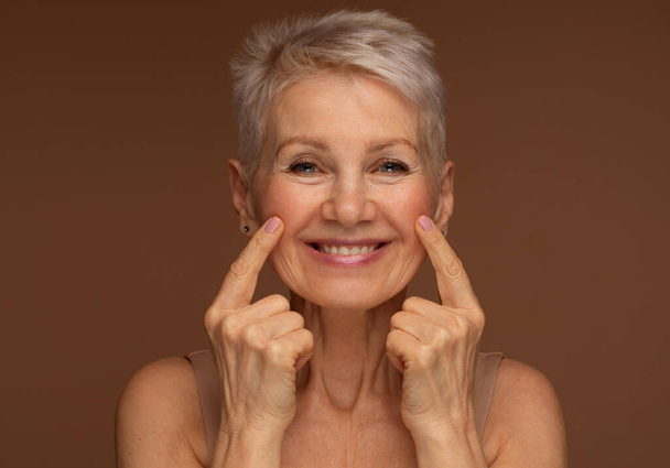 Portrait of beautiful senior woman touching her perfect skin and looking at camera. Closeup face of mature woman with wrinkles massaging face isolated over brown background. Aging process concept. - Фото, изображение