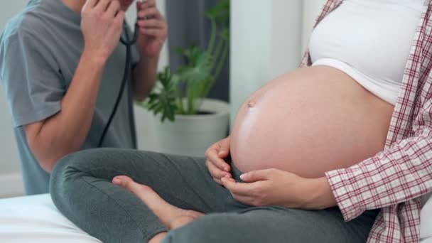 Fogantatott terhes nők és a férj. A férj sztetoszkóppal hallgatja a baba hangját. Hallgatni a meg nem született gyermek hangját, aki lehetséges rendellenességeket észlel. - Felvétel, videó