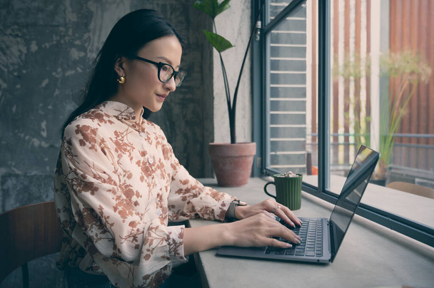 Bella cinese asiatica donna d'affari indossa gli occhiali, facendo lavoro freelance in caffè vicino alla finestra utilizzando il computer portatile. Lavoro online durante la pausa caffè. Digitando sulla tastiera. Giovani studenti studiano in caffetteria - Foto, immagini