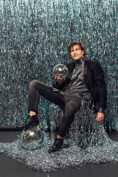 Tall attractive man sitting on the floor over the sparkle background and mirror disco ball holding glass of champagne. Party concept. - Фото, изображение