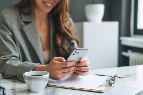 Crop zdjęcie młodej uśmiechniętej brunetki z długimi włosami w stylowym garniturze za pomocą telefonu komórkowego w rękach pracujących w jasnym nowoczesnym biurze, kobieta wysłać wiadomość przez smartfona - Zdjęcie, obraz