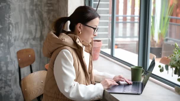 Hübsche chinesische asiatische Geschäftsfrau mit Brille, die mit Laptop im Café am Fenster arbeitet. In der Kaffeepause online arbeiten. Tippen auf der Tastatur. Junge Studentin studiert im Coffeeshop - Filmmaterial, Video