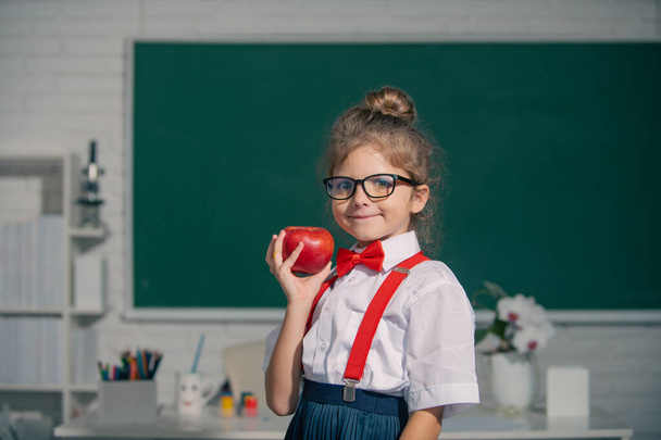 Schoolkind houdt appel met grappige uitdrukking tegen schoolbord. Onderwijs en kennis voor kinderen. - Foto, afbeelding