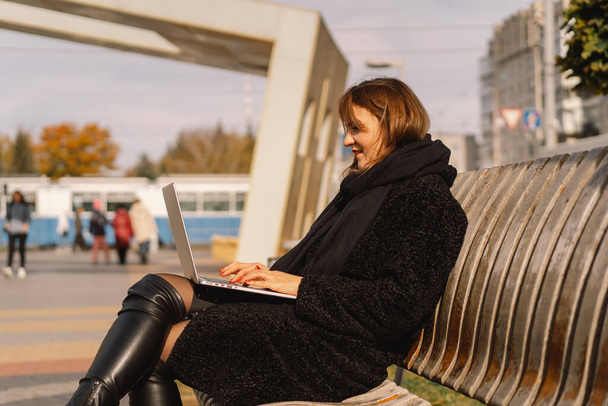 Μια γυναίκα κάθεται σε ένα παγκάκι στην πόλη, χρησιμοποιεί έναν υπολογιστή και μιλάει σε μια βιντεοκλήση. Ανεξάρτητη γυναίκα.  - Φωτογραφία, εικόνα