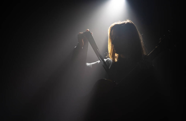 Meisje met een akoestische gitaar in het donker onder een lichtstraal. - Foto, afbeelding