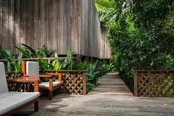 Кресло на деревянной тропинке, чтобы прогуляться до деревянного коттеджа на курорте с зеленой окружающей средой сада. Отдыхающий или летний отдых в тропической стране, Таиланде или Сиаме. - Фото, изображение