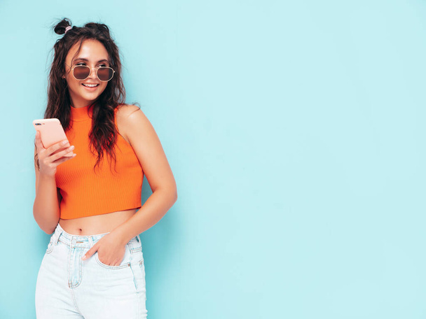 Young beautiful smiling female in trendy summer orange top and jeans clothes. Sexy carefree woman posing near blue wall in studio. Looking at smartphone screen. Model using cellphone apps - Photo, image