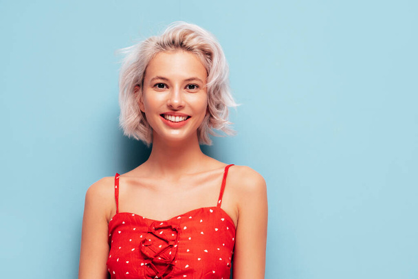 Portrait of young beautiful smiling female in trendy summer red dress. Sexy carefree woman posing near blue wall in studio. Positive blond model having fun and going crazy. Cheerful and happy - Φωτογραφία, εικόνα