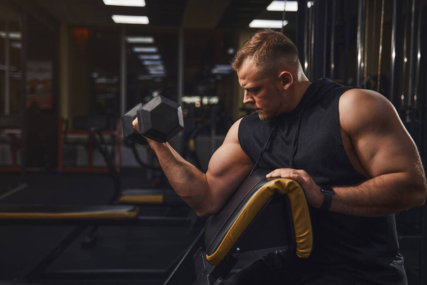 Hombre deportivo con grandes músculos y amplios trenes traseros en el gimnasio con pesas mientras balancea bíceps  - Foto, imagen