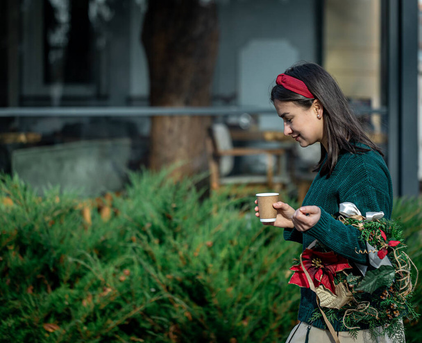 Νεαρή γυναίκα με Χριστουγεννιάτικο στεφάνι και ένα φλιτζάνι καφέ σε μια βόλτα στην πόλη. - Φωτογραφία, εικόνα
