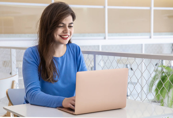 Hymyilevä nuori nainen sinisessä puserossa työskentelee kannettavassa kahvilassa. - Valokuva, kuva