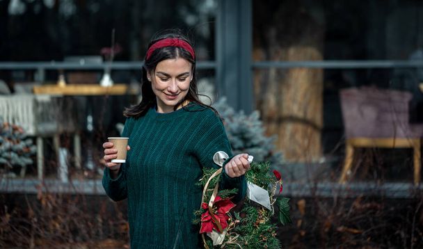 Młoda kobieta z świątecznym wieńcem i filiżanką kawy na spacerze po mieście. - Zdjęcie, obraz