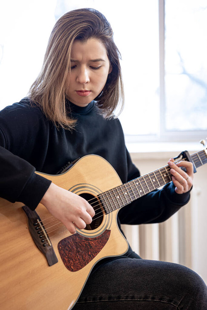 Serious young woman playing acoustic guitar at home. - Фото, изображение