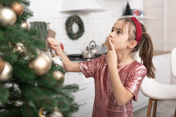 Ένα κοριτσάκι στολίζει ένα χριστουγεννιάτικο δέντρο, κρεμάει μπάλες. - Φωτογραφία, εικόνα