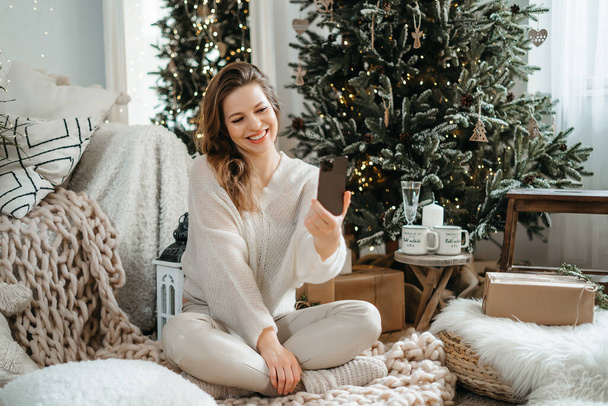 Задоволена молодою щасливою жінкою в теплому в'язаному светрі сидить під прикрашеним різдвяним деревом, веде розмову на мобільному телефоні з родичами або друзями, вітаючи найкращі побажання.. - Фото, зображення