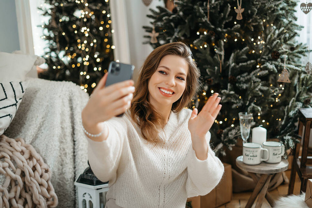 Überglückliche junge Europäerin in warmem Strickpullover sitzt unter geschmücktem Weihnachtsbaum, telefoniert mit Verwandten oder Freunden, gratuliert mit den besten Wünschen. - Foto, Bild