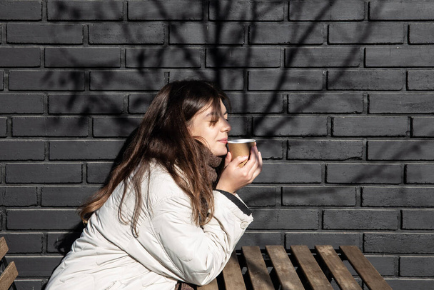 Jonge vrouw genieten van een drankje tegen de bakstenen muur van de coffeeshop buitenkant. - Foto, afbeelding