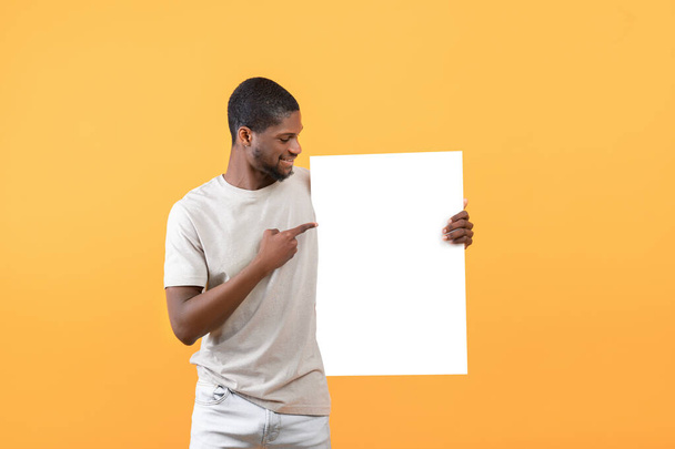 Рекламный щит. Портрет счастливого черного парня, указывающего на белый плакат с макетом, свободное пространство для вашего дизайна - Фото, изображение