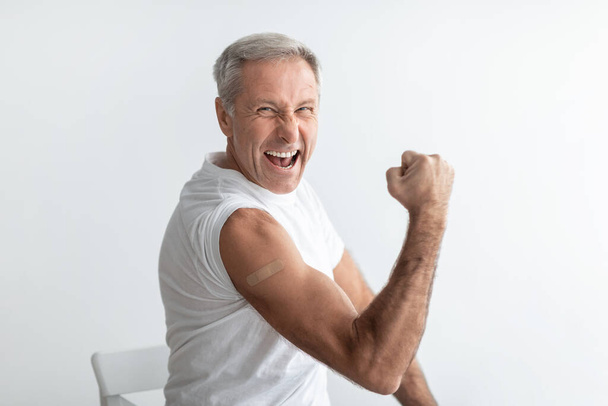 Alegre vacunado senior macho mostrando bíceps sobre gris fondo del estudio - Foto, imagen