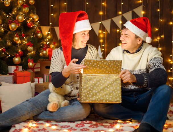 Προσωπογραφία ζευγαριού στη διακόσμηση της Πρωτοχρονιάς. Ανοίγουν ένα κουτί και διασκεδάζουν. Γιορτινά φώτα, δώρα και χριστουγεννιάτικο δέντρο στολισμένο με παιχνίδια. - Φωτογραφία, εικόνα