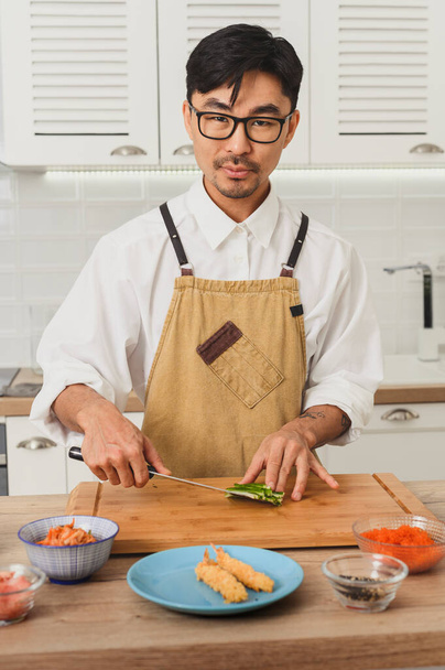 Japan voedsellevering concept. Vooraanzicht portret van Aziatische lachende chef-kok in uniform snijden komkommers voor sushi, kijken naar camera en glimlachen - Foto, afbeelding