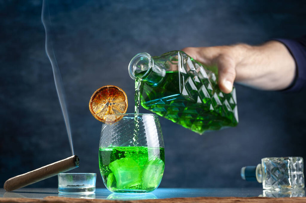 Uma vista de um decantador derramando uma bebida verde no copo com gelo e um charuto perto dele - Foto, Imagem