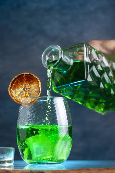 A vertical shot of a decanter pouring a green drink in the glass with ice and slices of lime - Foto, Imagem