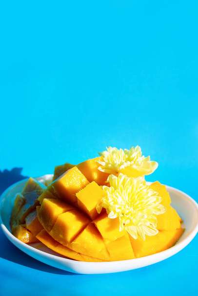 Красиво нарезанный манго лежит на тарелке в виде сердца на синем фоне. Экзотические фрукты, украшенные цветками хризантемы - Фото, изображение
