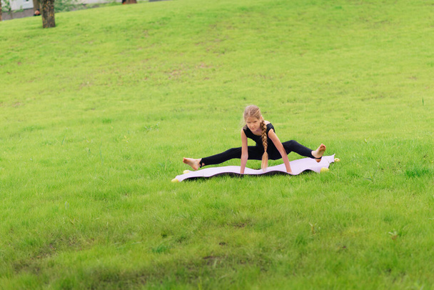 slim girl in a black gymnastic uniform performs gymnastic exercises or pilates on the green grass in the park. Professional gymnast during training - Foto, Bild