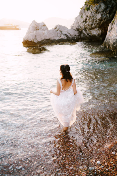 Noiva caminha no mar, segurando a bainha de seu vestido. Visão traseira - Foto, Imagem
