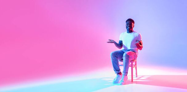 Full length of happy black guy sitting on chair, offering free space for your advertisement in neon light, banner - Foto, imagen