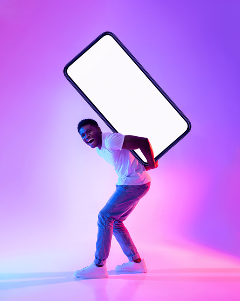 Young black guy carrying big heavy smartphone with empty screen on his back in neon light, full length. Mockup - Foto, Imagen