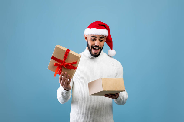 Hombre árabe alegre esperando la Navidad, abriendo la caja de regalo envuelta y mirando hacia el interior, posando sobre fondo azul - Foto, Imagen
