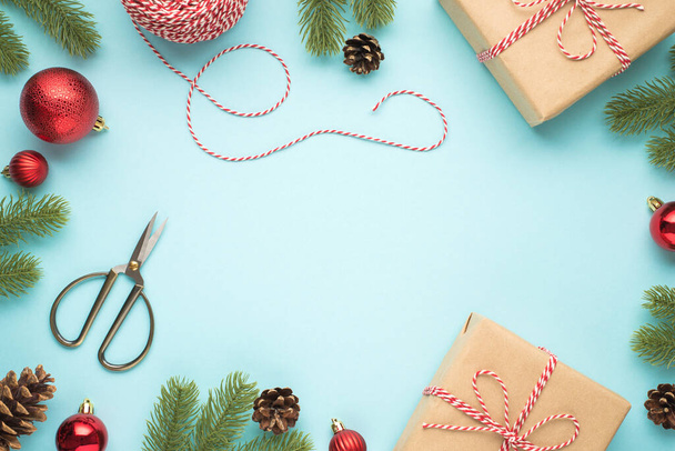 Nahoru pohled fotografie borovice kužely červené vánoční stromeček koule nůžky cívka ozdobného lana a dva řemeslné papírové dárkové krabice na izolované pastelové modré pozadí s copyspace uprostřed - Fotografie, Obrázek