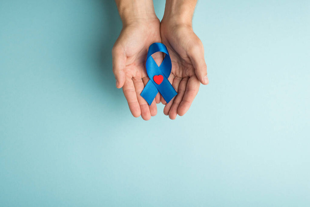 First person top view photo of male hands demonstrating blue ribbon with small red heart in palms symbol of prostate cancer awareness on isolated pastel blue background with empty space - Foto, Imagen
