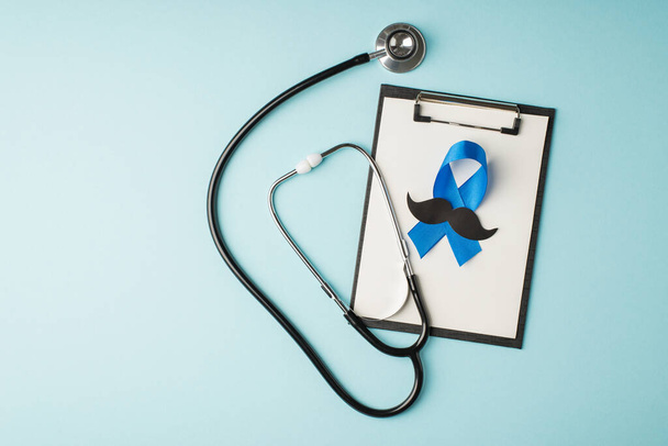 Top view φωτογραφία του προχείρου στηθοσκόπιο και μπλε κορδέλα με μουστάκι σιλουέτα σύμβολο της ευαισθητοποίησης του καρκίνου του προστάτη σε απομονωμένο παστέλ μπλε φόντο με copyspace - Φωτογραφία, εικόνα