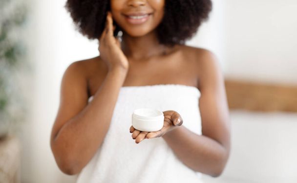Beauty skin procedure concept. Closeup view of young black woman applying facial cream from jar at home, selective focus - 写真・画像