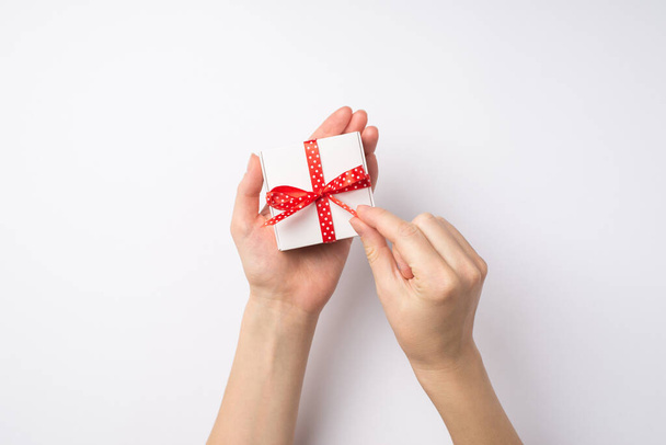 Première personne vue du dessus photo des mains de la jeune femme déballer petite boîte-cadeau blanche avec ruban rouge pointillé arc sur fond blanc isolé - Photo, image
