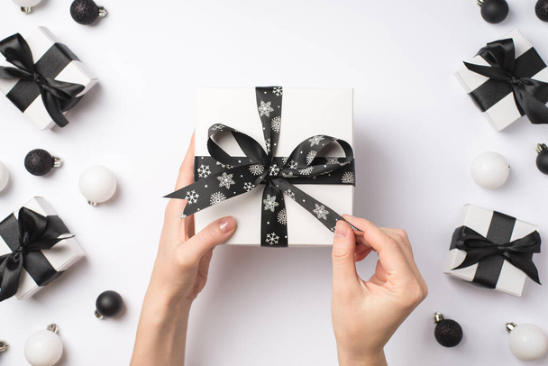 First person top view photo of young girl's hands unwrapping white giftbox with black ribbon bow over white and black christmas decorations presents and balls on isolated white background - Photo, Image