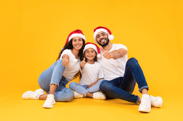 Glückliche arabische Familie in Weihnachtsmützen mit Daumen hoch, gelbem Hintergrund - Foto, Bild